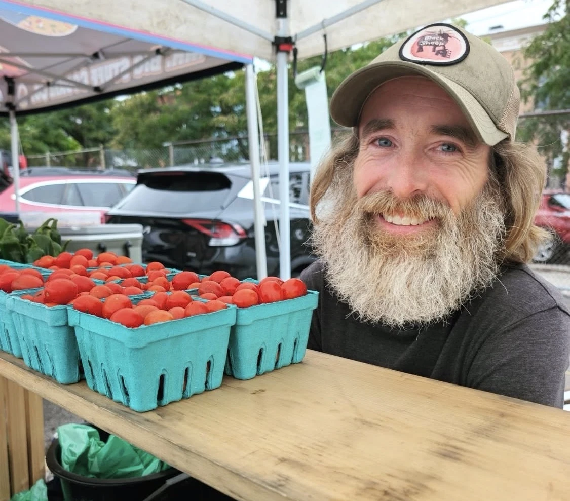 A smiling grower beside baskets of tomatoes at the market