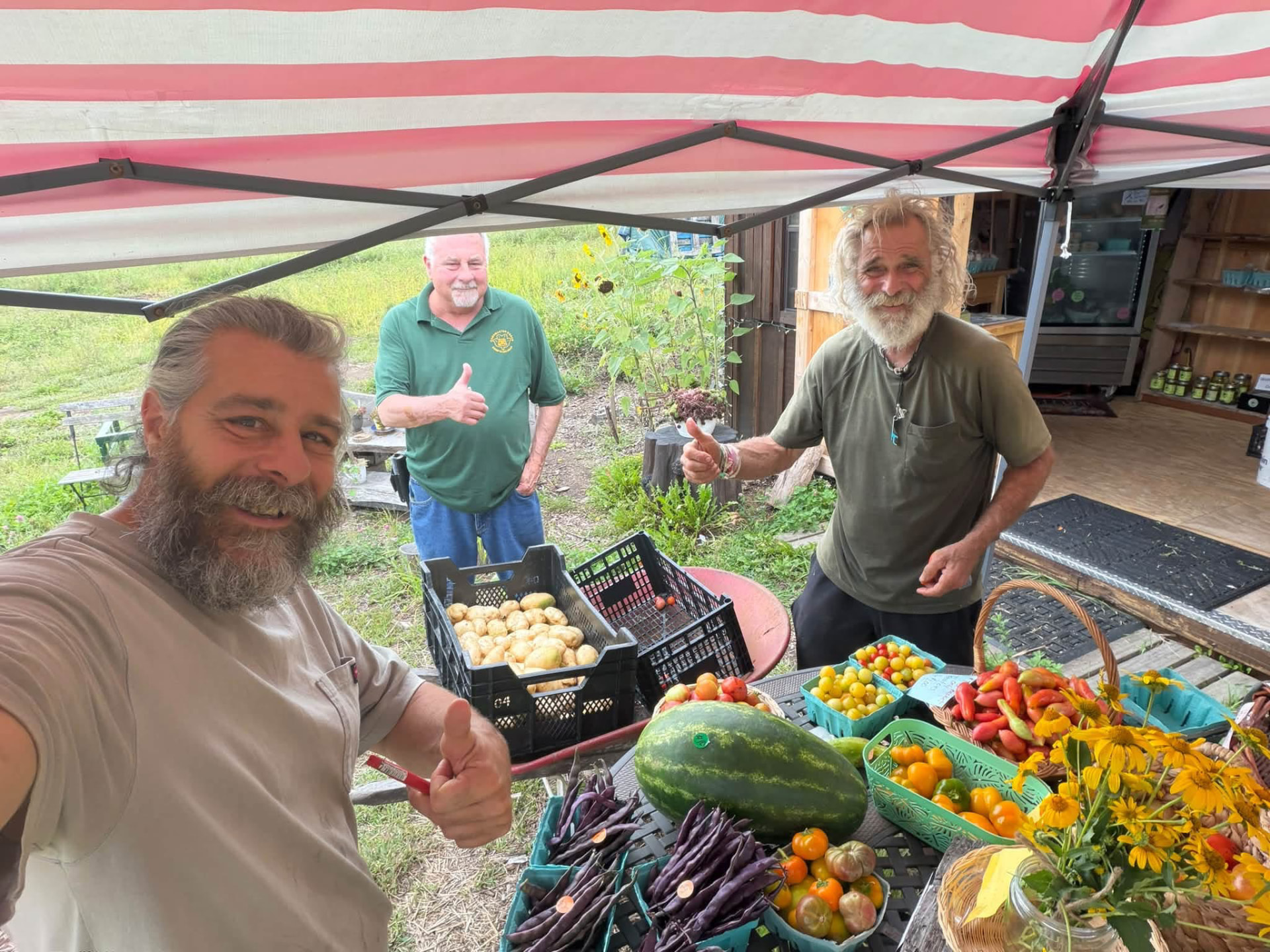Three smiling visitors giving a thumbs-up beside a colorful summer harvest at the farm stand